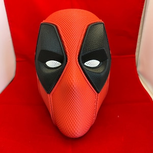 May include: A full-head mask featuring a textured red exterior with a fine grid pattern. The mask includes large, black, leather-like eye patches surrounding white, mesh-covered eye openings, creating a distinct character-inspired appearance.