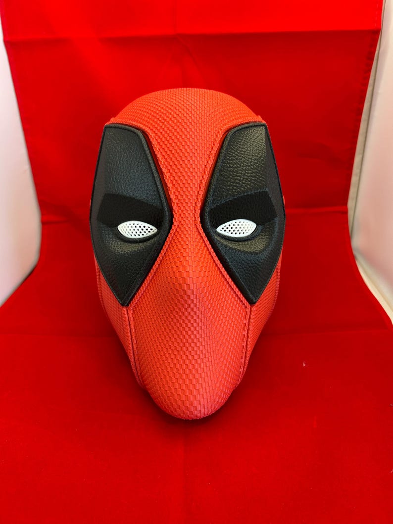 May include: A full-head mask featuring a textured red exterior with a fine grid pattern. The mask includes large, black, leather-like eye patches surrounding white, mesh-covered eye openings, creating a distinct character-inspired appearance.