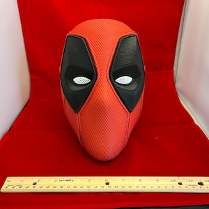 May include: A red and black textured mask with white mesh eye coverings. A wooden ruler is placed in front of the mask, showing a length of 12 inches. The mask features a distinct cross-stitched seam down the center and black eye patches.