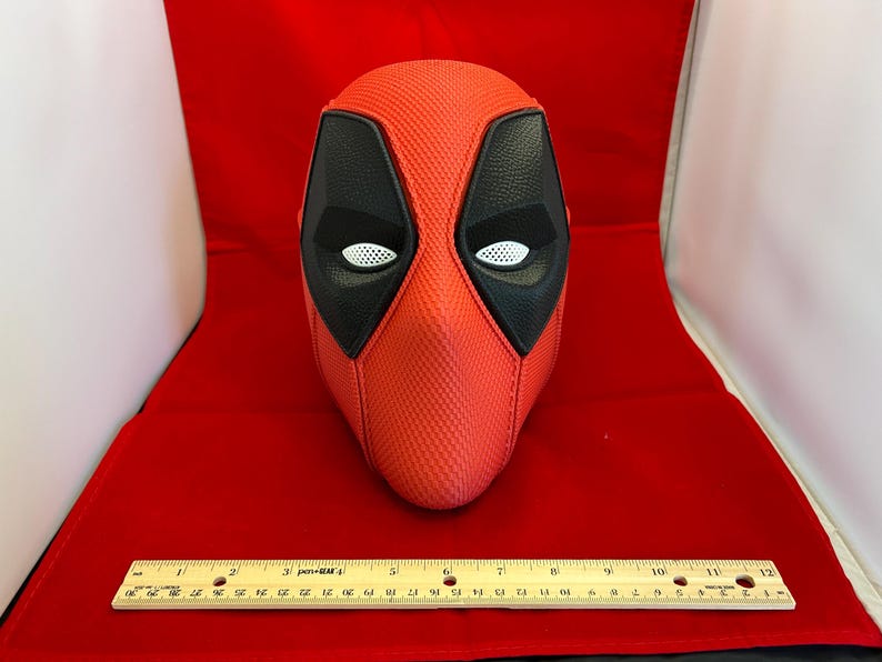 May include: A red and black textured mask with white mesh eye coverings. A wooden ruler is placed in front of the mask, showing a length of 12 inches. The mask features a distinct cross-stitched seam down the center and black eye patches.