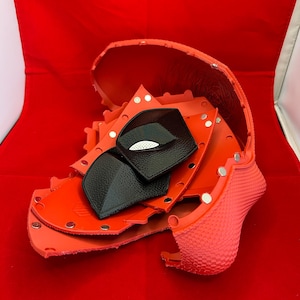 May include: A disassembled red helmet shell with a textured exterior and smooth interior, featuring multiple silver metal snaps along the edges. Two black, textured, diamond-shaped eye pieces with white mesh accents are placed inside the shell.