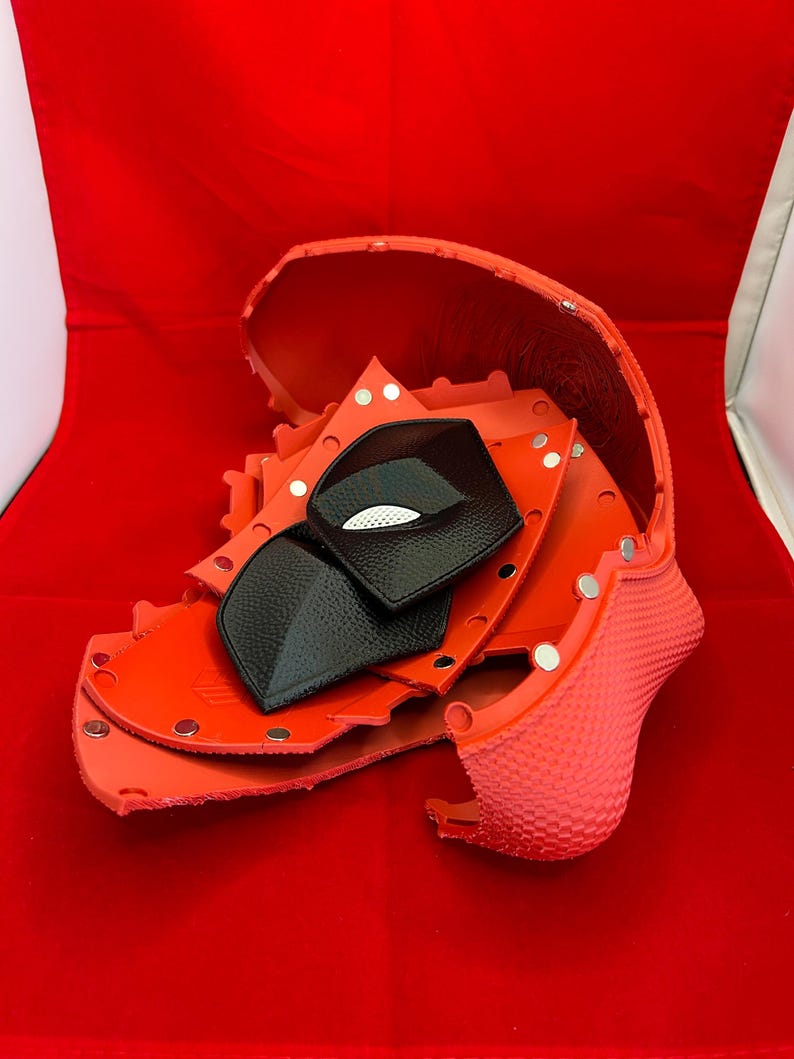 May include: A disassembled red helmet shell with a textured exterior and smooth interior, featuring multiple silver metal snaps along the edges. Two black, textured, diamond-shaped eye pieces with white mesh accents are placed inside the shell.