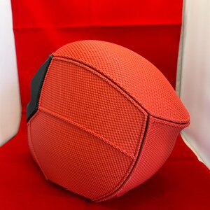 May include: A rounded, structured object featuring a vibrant orange, textured, grid-like fabric surface. The item is composed of multiple panels joined by visible seams, with a solid black accent piece visible on the left side of the curved form.