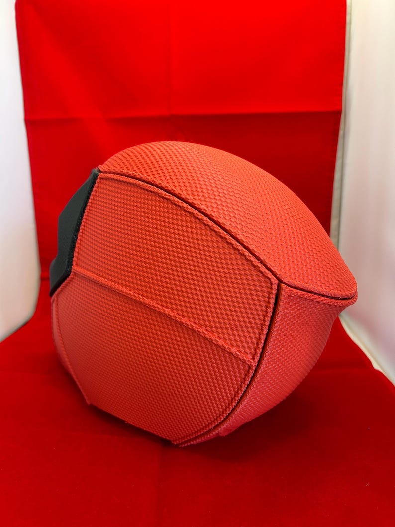 May include: A rounded, structured object featuring a vibrant orange, textured, grid-like fabric surface. The item is composed of multiple panels joined by visible seams, with a solid black accent piece visible on the left side of the curved form.