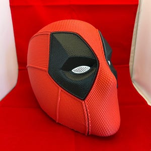May include: A full-head mask featuring a textured red surface with prominent black eye patches. The eye areas contain white mesh inserts. The mask has a distinct seam running vertically down the center and across the sides, creating a structured appearance.