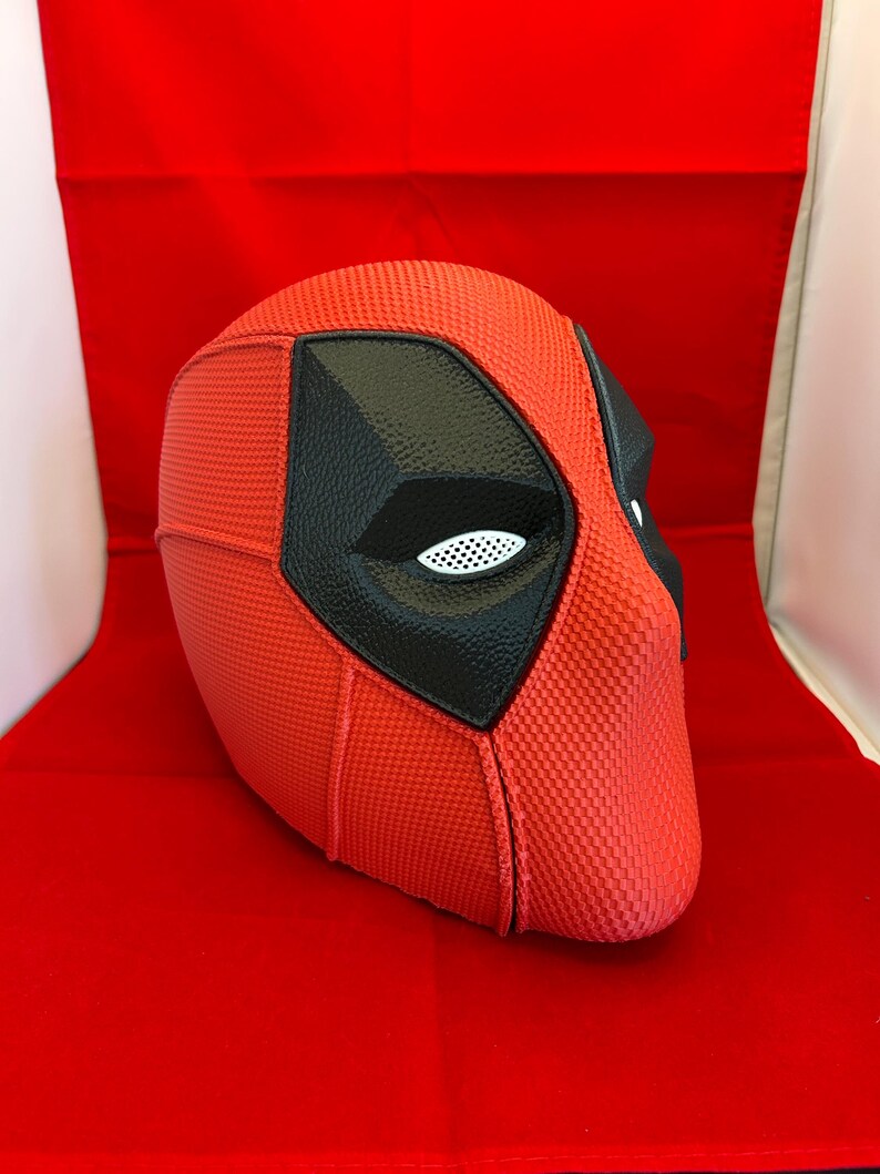 May include: A full-head mask featuring a textured red surface with prominent black eye patches. The eye areas contain white mesh inserts. The mask has a distinct seam running vertically down the center and across the sides, creating a structured appearance.