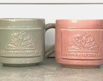 May include: Two ceramic mugs, one sage green and one dusty pink, each featuring a raised embossed design of an open book with flowers emerging from the pages and the text "JUST ONE MORE CHAPTER..." centered below the book illustration.