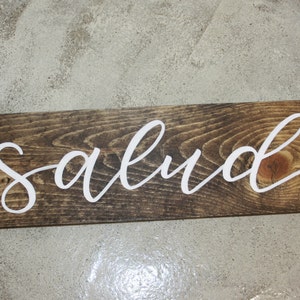 Puede incluir: Un letrero rectangular de madera con la palabra "salud" en letras cursivas blancas. La madera tiene una mancha marr&oacute;n oscura y la veta es visible. El letrero es probablemente para la decoraci&oacute;n del hogar.