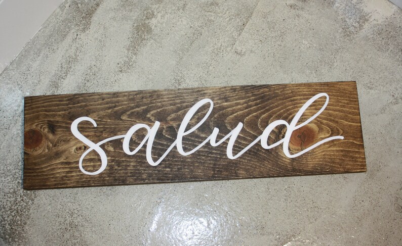 Puede incluir: Un letrero rectangular de madera con la palabra "salud" en letras cursivas blancas. La madera tiene una mancha marr&oacute;n oscura y la veta es visible. El letrero es probablemente para la decoraci&oacute;n del hogar.
