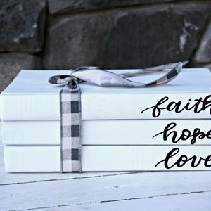 May include: Three white books stacked on top of each other. The books are tied together with a black and white checked ribbon. The top book has the word "faith" written on it in black cursive. The middle book has the word "hope" written on it in black cursive. The bottom book has the word "love" written on it in black cursive.