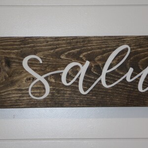 Puede incluir: Un letrero rectangular de madera con la palabra "salva" en letras cursivas blancas. La madera tiene un tinte marr&oacute;n oscuro, y el letrero est&aacute; montado en una pared blanca con paneles horizontales. El letrero es un art&iacute;culo decorativo.