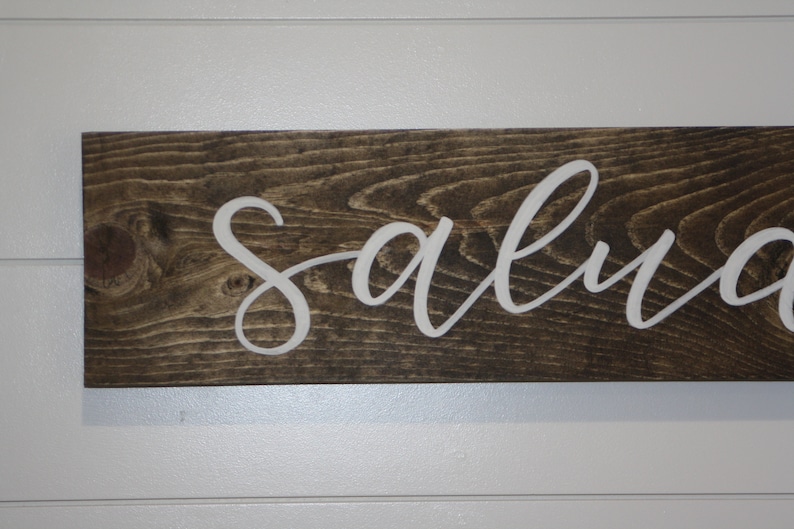 Puede incluir: Un letrero rectangular de madera con la palabra "salva" en letras cursivas blancas. La madera tiene un tinte marr&oacute;n oscuro, y el letrero est&aacute; montado en una pared blanca con paneles horizontales. El letrero es un art&iacute;culo decorativo.