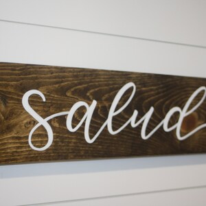Puede incluir: Un letrero rectangular de madera con la palabra "Salud" en letras blancas cursivas. La madera tiene una mancha marr&oacute;n oscura y el letrero est&aacute; montado en una pared blanca con paneles horizontales. Es un art&iacute;culo decorativo.