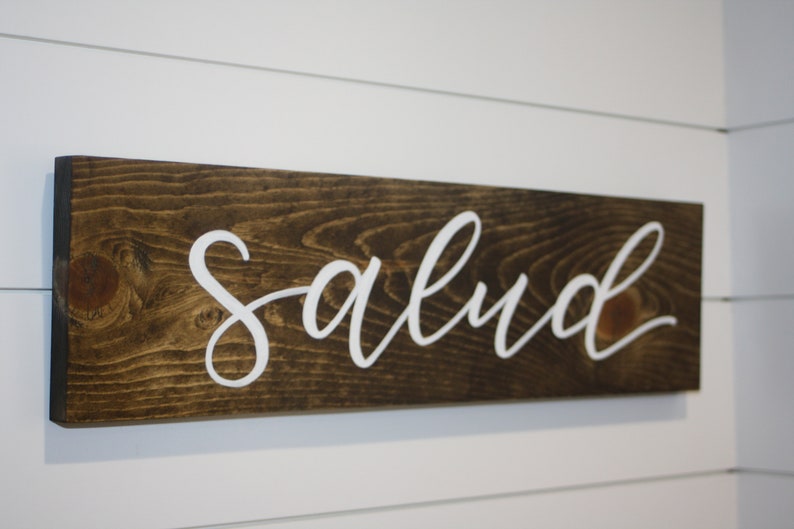 Puede incluir: Un letrero rectangular de madera con la palabra "Salud" en letras blancas cursivas. La madera tiene una mancha marr&oacute;n oscura y el letrero est&aacute; montado en una pared blanca con paneles horizontales. Es un art&iacute;culo decorativo.