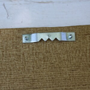 Puede incluir: Primer plano de la parte posterior de un marco de madera con un colgador de metal en forma de sierra. El colgador est&aacute; asegurado con tornillos y tiene una serie de dientes angulados para colgar. La madera tiene un aspecto marr&oacute;n texturizado.