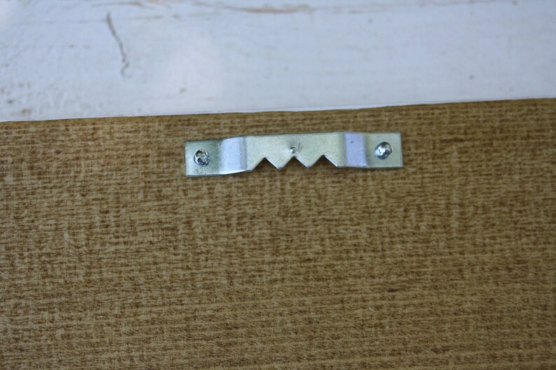 Puede incluir: Primer plano de la parte posterior de un marco de madera con un colgador de metal en forma de sierra. El colgador est&aacute; asegurado con tornillos y tiene una serie de dientes angulados para colgar. La madera tiene un aspecto marr&oacute;n texturizado.