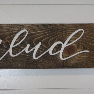 Puede incluir: Un letrero de madera con la palabra "abud" en letras blancas cursivas. El letrero est&aacute; hecho de madera te&ntilde;ida oscura con vetas visibles. El letrero est&aacute; enmarcado con madera pintada de blanco.