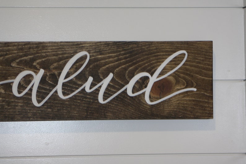 Puede incluir: Un letrero de madera con la palabra "abud" en letras blancas cursivas. El letrero est&aacute; hecho de madera te&ntilde;ida oscura con vetas visibles. El letrero est&aacute; enmarcado con madera pintada de blanco.