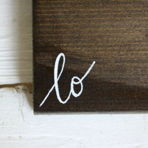 Puede incluir: Primer plano de un letrero de madera marr&oacute;n oscuro con la palabra "lo" escrita en escritura blanca en la esquina inferior izquierda. El letrero est&aacute; sobre un fondo blanco texturizado.