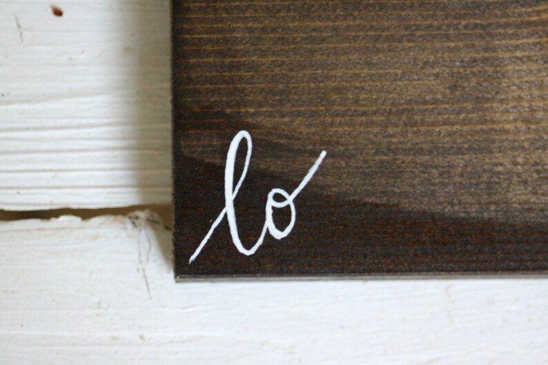 Puede incluir: Primer plano de un letrero de madera marr&oacute;n oscuro con la palabra "lo" escrita en escritura blanca en la esquina inferior izquierda. El letrero est&aacute; sobre un fondo blanco texturizado.