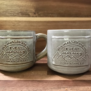 May include: Two ceramic mugs with a light gray glaze. Each mug features a rectangular panel with the text "His mercies are new every morning" and "Lamentations 3:23" in a dark gray color. The mugs have curved handles.
