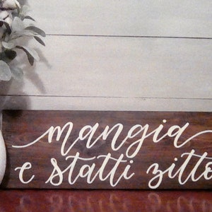May include: A wooden sign with the Italian phrase "mangia e stati zitto" written in white cursive script.