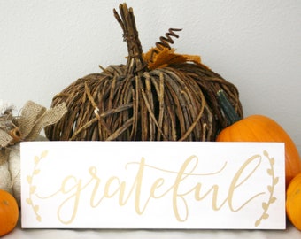 May include: A white rectangular sign with the word "grateful" in gold script, flanked by leaf designs. A woven pumpkin and several orange pumpkins are in the background, creating a fall-themed display.