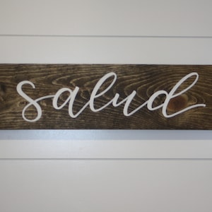 Puede incluir: Un letrero rectangular de madera con la palabra "salud" en letras cursivas blancas. El letrero es de madera marr&oacute;n oscuro con vetas visibles y est&aacute; montado en una pared blanca.