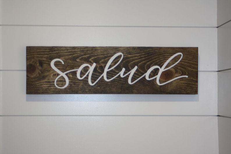 Puede incluir: Un letrero rectangular de madera con la palabra "salud" en letras cursivas blancas. El letrero es de madera marr&oacute;n oscuro con vetas visibles y est&aacute; montado en una pared blanca.