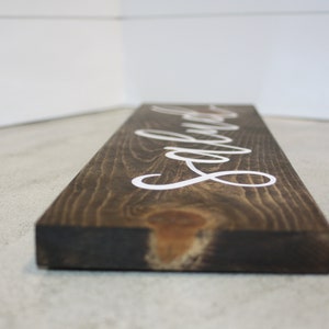 Puede incluir: Un letrero rectangular de madera te&ntilde;ida oscura con la palabra "Laundry" en letras cursivas blancas. La veta de la madera es visible y el letrero se ve desde un &aacute;ngulo bajo.