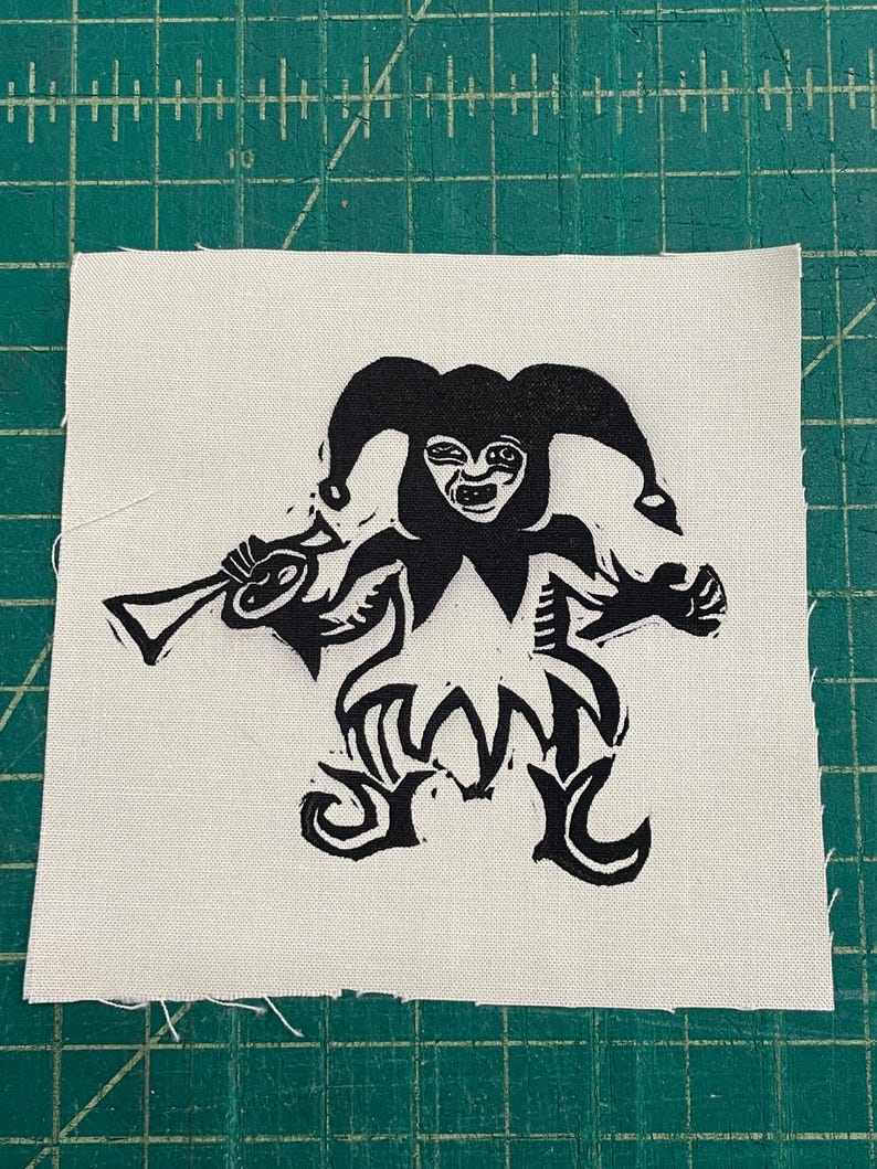 May include: A black and white patch featuring a jester figure holding a horn. The jester has a smiling face and is wearing a hat with three points. The patch is on a white square of fabric.