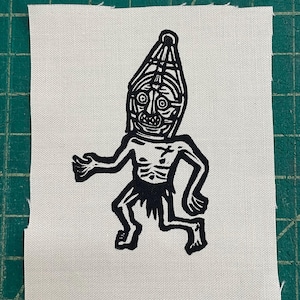 Puede incluir: Un parche en blanco y negro que presenta una figura estilizada con una cara similar a una máscara y un tocado. La figura está representada con un estilo minimalista, con un contorno simple y líneas audaces. El parche está hecho de tela blanca.