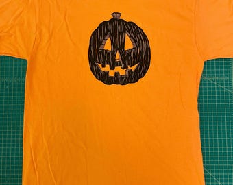 May include: A bright orange short-sleeve t-shirt featuring a centered black graphic print of a carved jack-o'-lantern pumpkin. The pumpkin design has a textured, hand-drawn appearance with triangular eyes and a jagged, toothy grin on the front.