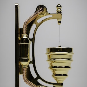 May include: A gold-toned metal wall-mounted soap dispenser with a decorative design. The dispenser has a coiled copper pipe and a round, tiered soap dish.