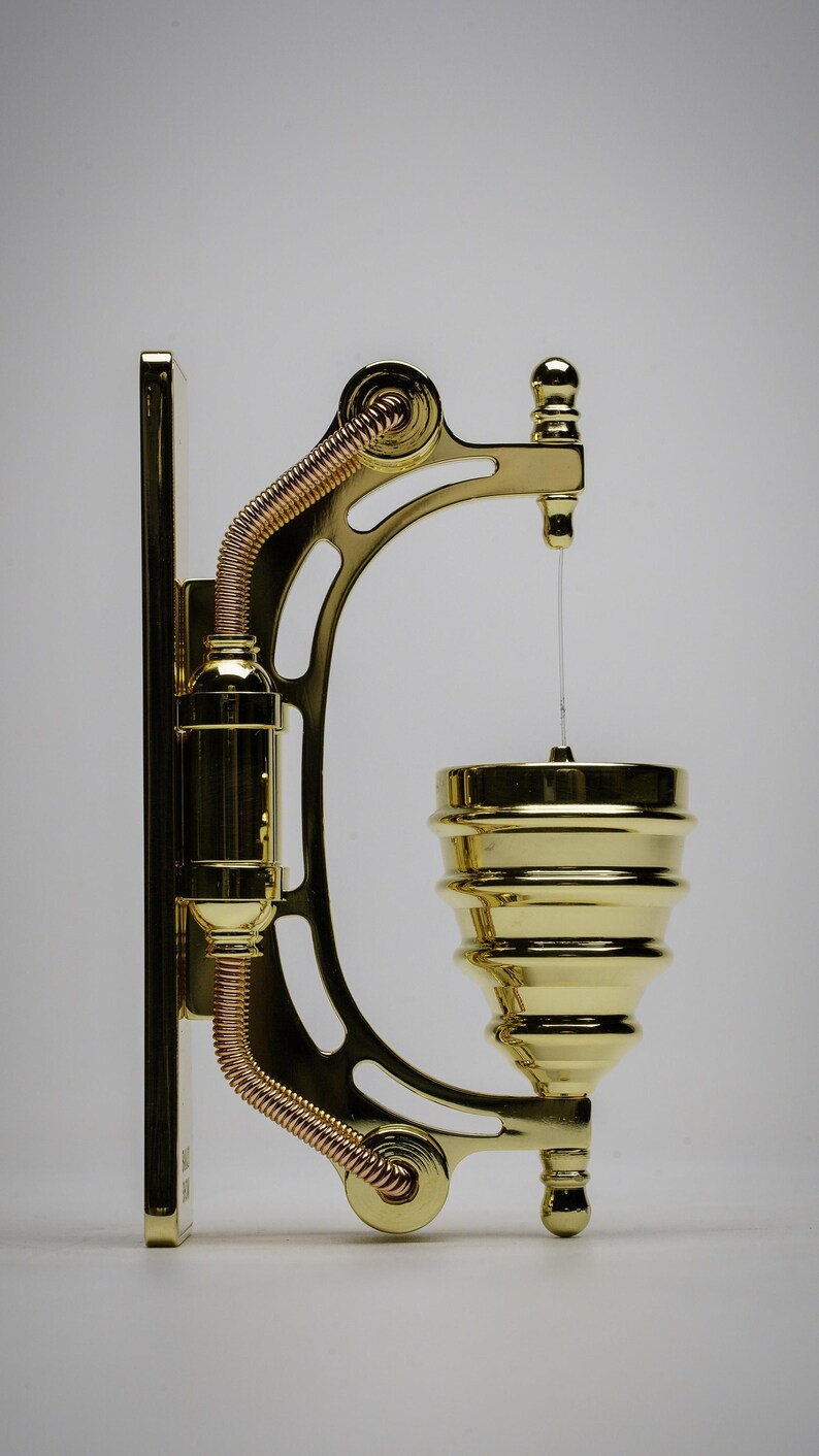 May include: A gold-toned metal wall-mounted soap dispenser with a decorative design. The dispenser has a coiled copper pipe and a round, tiered soap dish.