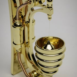 May include: A gold-toned metal wall-mounted steampunk-style water dispenser with a copper-colored pipe and a small, round, gold-toned bowl at the bottom.