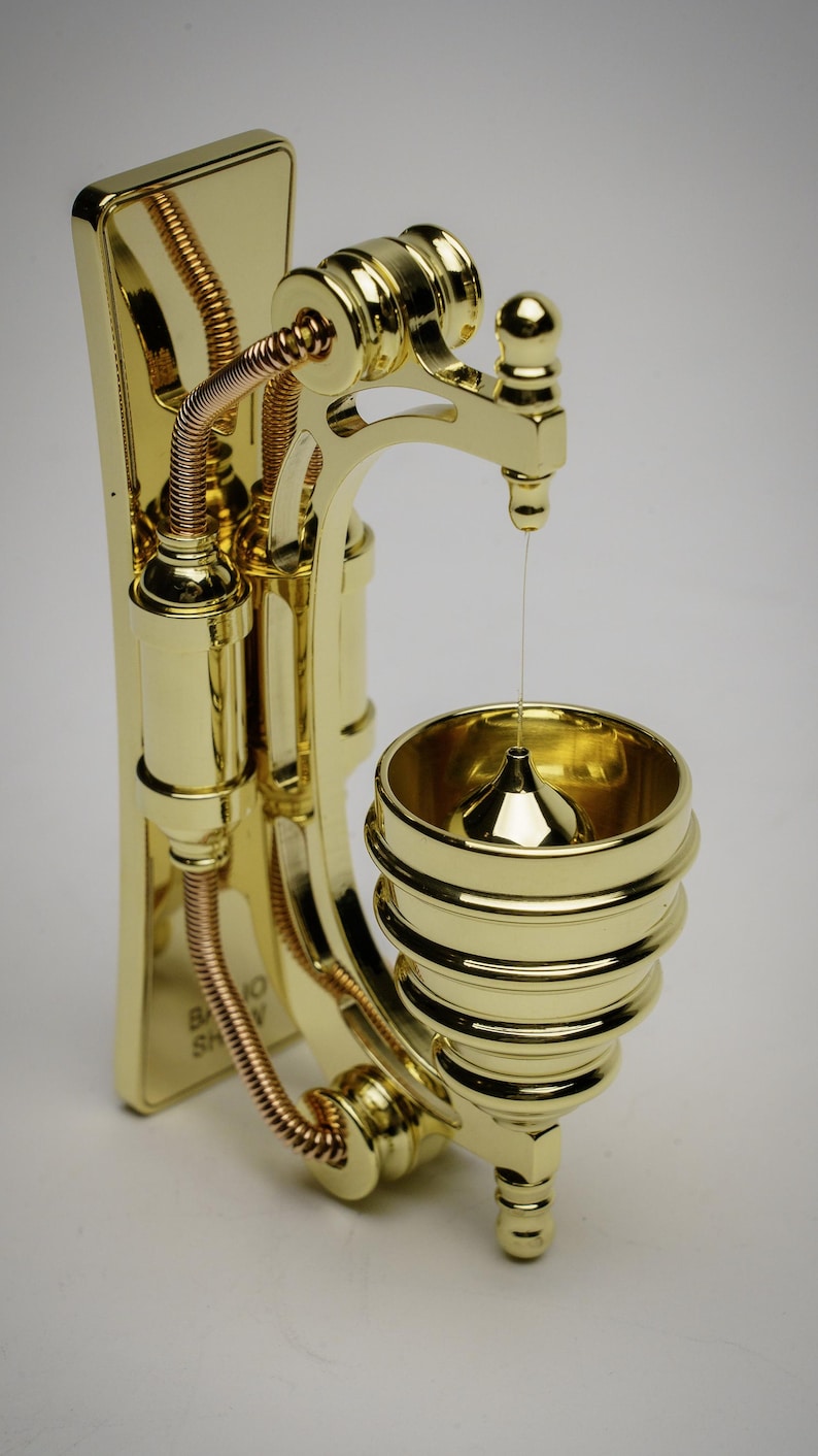 May include: A gold-toned metal wall-mounted steampunk-style water dispenser with a copper-colored pipe and a small, round, gold-toned bowl at the bottom.