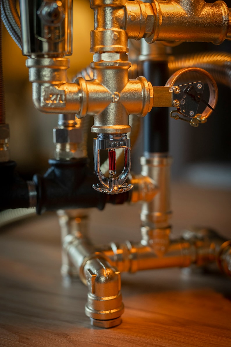 May include: A gold metal sprinkler head with a red heat sensor attached to a pipe system. The sprinkler head is mounted on a gold metal pipe system.