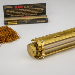 May include: A gold metal cigarette roller with a clear plastic tube. The roller has a RAW logo on it. There is a pile of tobacco and a pack of RAW Classic rolling papers in the background.