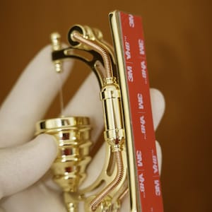 May include: A gold-colored metal object with a decorative design. It has a curved shape and appears to be a decorative piece or a functional tool. The object has a series of small, round details and a thin, copper-colored wire running along its length. The object is held in a gloved hand. The object has a red adhesive tape with the text "3M VHB&trade;" printed on it.