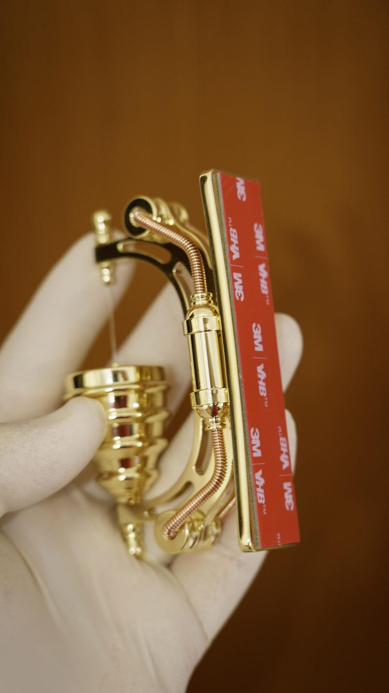 May include: A gold-colored metal object with a decorative design. It has a curved shape and appears to be a decorative piece or a functional tool. The object has a series of small, round details and a thin, copper-colored wire running along its length. The object is held in a gloved hand. The object has a red adhesive tape with the text "3M VHB&trade;" printed on it.
