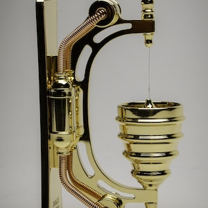 May include: A gold-toned metal wall-mounted soap dispenser with a decorative design. The dispenser has a small, round, gold-toned soap dish with a thin, clear line of soap hanging from it. The dispenser is labeled "BANJO SHOW".