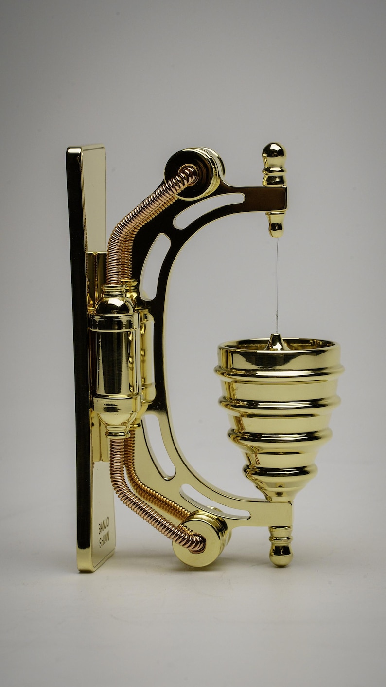 May include: A gold-toned metal wall-mounted soap dispenser with a decorative design. The dispenser has a small, round, gold-toned soap dish with a thin, clear line of soap hanging from it. The dispenser is labeled "BANJO SHOW".