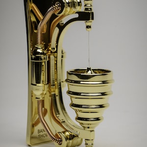 May include: A gold-colored metal wall-mounted spinning top with a decorative design. The top is suspended by a thin string and has a base with the text "BAN SHOW" on it.