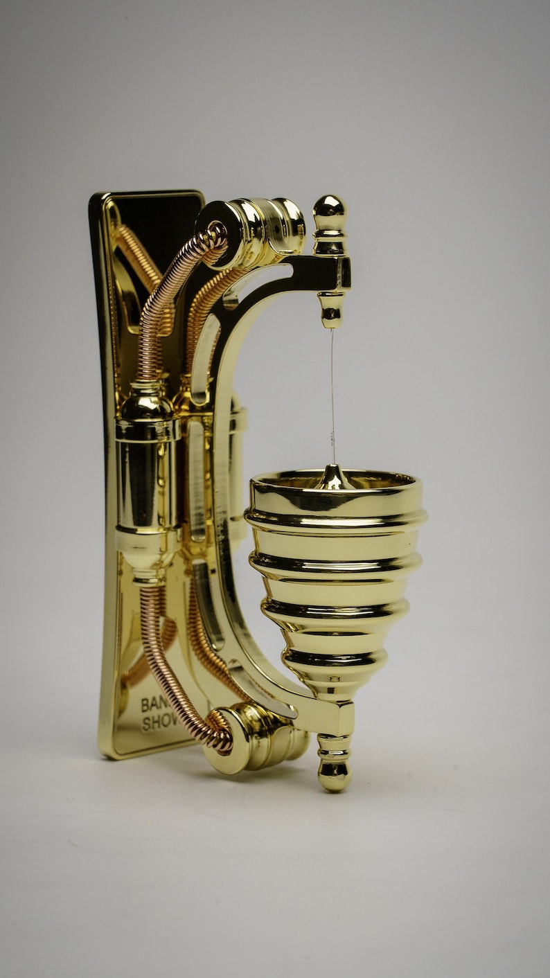 May include: A gold-colored metal wall-mounted spinning top with a decorative design. The top is suspended by a thin string and has a base with the text "BAN SHOW" on it.