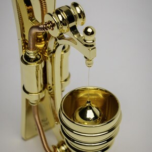 May include: A gold-toned metal wall-mounted oil dispenser with a decorative, ornate design. The dispenser has a spout that drips oil into a small, round container below.