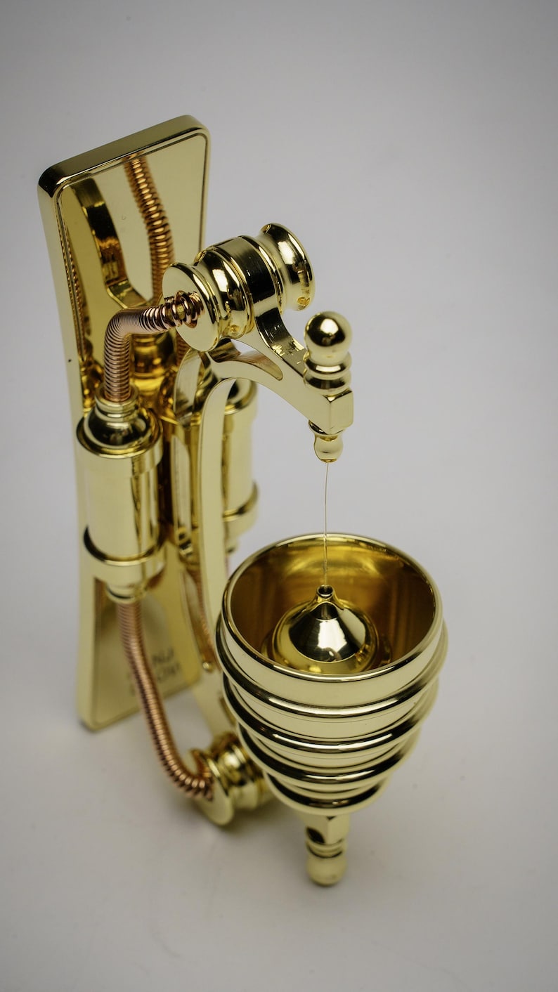 May include: A gold-toned metal wall-mounted oil dispenser with a decorative, ornate design. The dispenser has a spout that drips oil into a small, round container below.