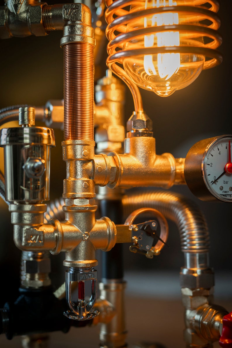 May include: A steampunk-style lamp made from gold-toned pipes and fittings. The lamp features a clear glass globe with a copper coil inside, a pressure gauge labeled "0 bar" and "FIMET 1/2 WATTS", and a sprinkler head.