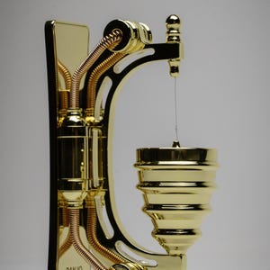 May include: A gold-colored metal wall-mounted banjo stand with a decorative design. The stand has a curved arm with a small cup at the end. The stand has the words "Banjo Show" printed on the side.