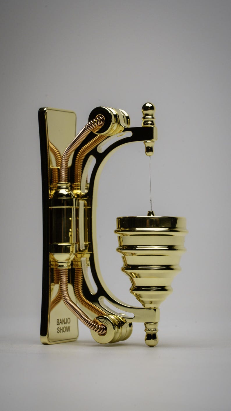 May include: A gold-colored metal wall-mounted banjo stand with a decorative design. The stand has a curved arm with a small cup at the end. The stand has the words "Banjo Show" printed on the side.